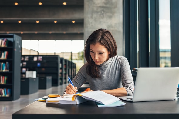 Imagem de uma mulher estudando em frente à um computador e fazendo anotações.